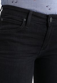 Jeans de mezclilla negra con diseño de cinco bolsillos, que presentan una cinturilla plana, botón de metal y un sutil detalle de costura en la parte frontal.