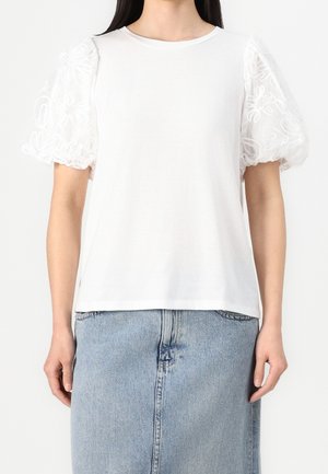 Mujer que lleva una blusa blanca con mangas abullonadas transparentes bordadas y una falda vaquera azul claro, de pie frente a un fondo liso.