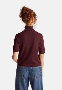 Frau mit lockigem, rotem Haar, die einen kurzärmeligen burgunderfarbenen Rollkragenpullover und blaue Jeans trägt. Sie steht mit dem Rücken zu einer schlichten Kulisse.