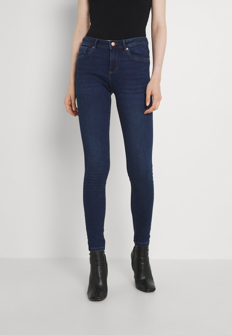 Femme portant un jean skinny bleu foncé et des bottines noires, debout devant un fond clair uni.
