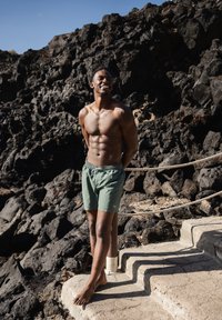 Un maillot de bain vert avec une taille élastique et un cordon de serrage, porté par une personne debout sur des marches en pierre à côté de rochers volcaniques foncés.