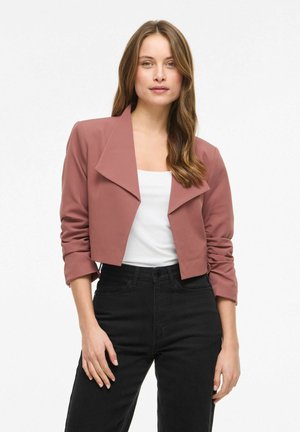 Femme portant un blazer rose poussière court par-dessus un haut blanc et un jean noir taille haute, debout devant un fond blanc.