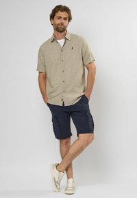 Beige kortærmet skjorte med knapper og krave samt et grønt logo, parret med marineblå cargo shorts med sidelommer og en afslappet pasform.