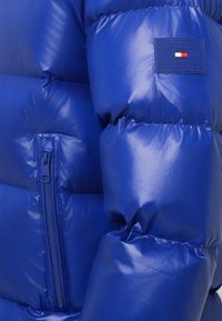 Niebieska kurtka puchowa o błyszczącym wykończeniu, z pikowanym wzorem, kieszenią na zamek błyskawiczny oraz naszywką z logo Tommy Hilfiger na klatce piersiowej.