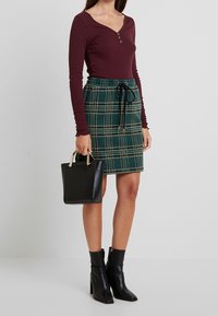 Kvinna klädd i en vinröd långärmad tröja, grön rutmönstrad kjol med snörning, svarta ankelboots, hållande en liten svart handväska med guldfärgad handtag.