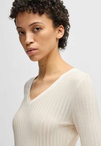 Light cream long-sleeve top with a V-neckline, featuring a textured ribbed pattern and a snug fit. Soft fabric, smooth finish, and minimal detailing.