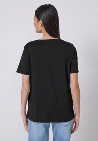Kvinde med langt m�ørkt hår iført en ensfarvet sort kortærmet skjorte og lyseblå jeans, stående med ryggen vendt mod en ensfarvet baggrund.