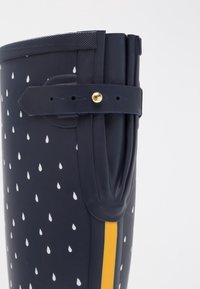 Botte de pluie bleu marine avec motif de gouttes de pluie blanches, rayure verticale jaune et sangle ajustable avec un bouton doré sur le côté.