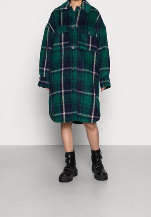 Robe-shirt oversize à carreaux verts et bleu marine en tissu doux, avec un col, deux poches avant et une fermeture à boutons. Portée avec des bottines noires.