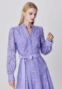 Robe à motif floral lilas avec un col, des manches longues et une taille ceinturée. Présente des détails de boutons sur le devant et un tissu texturé.
