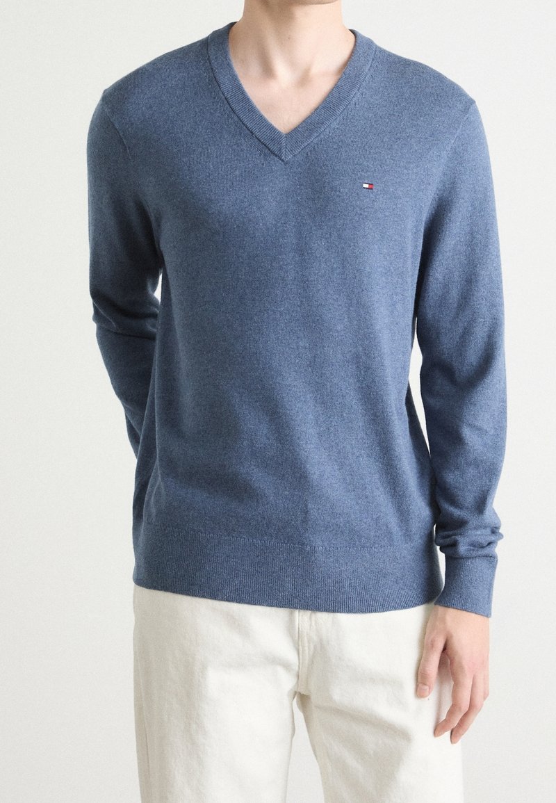 Homme portant un pull bleu à col en V manches longues avec un petit logo sur la poitrine et un pantalon écru sur un fond uni.