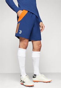 Pantaloni sportivi blu con accenti arancioni, caratterizzati da tre strisce e un logo. Realizzati in un materiale leggero, progettati per l'abbigliamento atletico.