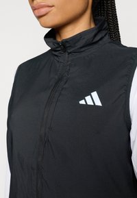 Svart vest med glidelås og høy krage, laget av glatt, lett materiale, med hvit Adidas-logo og kontrasterende hvite ermedetaljer.