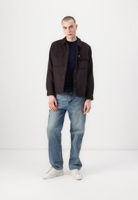 Chemise noire à bouton avec deux poches poitrine, associée à un jean bleu déchiré et des baskets blanches. Design simple, coupe décontractée.