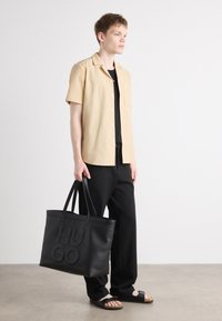 Beige Kurzarmhemd, schwarzes T-Shirt darunter, schwarze Hose, schwarze Tote-Tasche mit geprägtem "HUGO" und schwarze Sandalen mit einem Riemen.