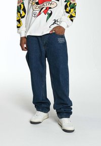 Baggy dark blue denim jeans with a white long-sleeve shirt featuring a graphic design. Shoes are white with black accents.