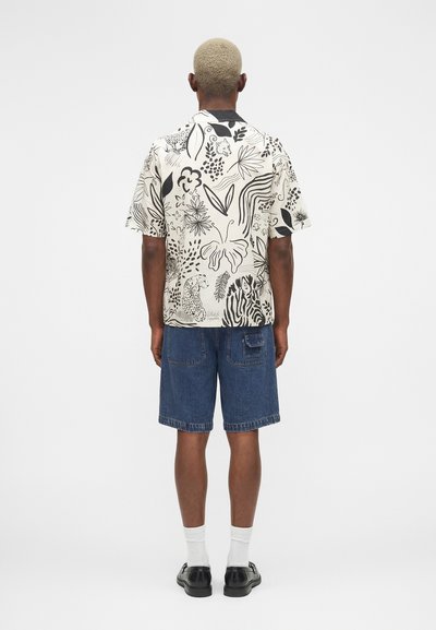 Άτομο με κοντά ξανθά μαλλιά που φοράει μαύρο-άσπρο πουκάμισο με μοτίβο και κοντά μανίκια, τζιν σορτς, λευκές κάλτσες και μαύρα παπούτσια, στέκεται με την πλάτη γυρισμένη.