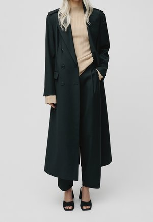 Kobieta o długich blond włosach, ubrana w czarny długi płaszcz, czarne szerokie spodnie, beżowy prążkowany golf i czarne czółenka z odkrytymi palcami.