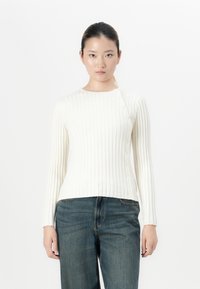 Vrouw in een witte ribbeltrui met knopen aan de zijkant en blauwe jeans, staande tegen een simpele witte achtergrond.