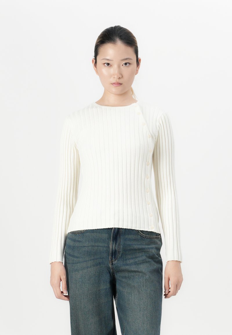 Vrouw in een witte ribbeltrui met knopen aan de zijkant en blauwe jeans, staande tegen een simpele witte achtergrond.