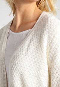 Bílý pletený kardigan s diamantovým vzorem, otevřený vpředu a měkkou texturou. Nesený přes jednoduché bílé tričko.