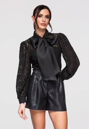 Femme portant un chemisier en dentelle noire avec un grand nœud en satin et un short en cuir noir taille haute, posant devant un fond gris clair.