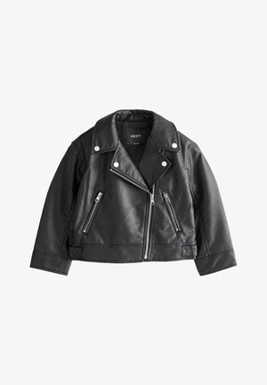 Junges Mädchen steht mit den Händen in den Taschen, trägt eine schwarze Lederjacke, eine weiße Bluse, Jeans mit weitem Bein, silberne Ballerinas und eine Kappe mit Leopardenmuster.