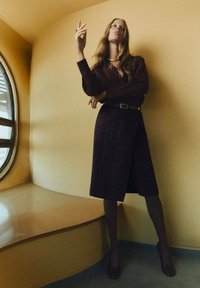 Burgundy blouse tucked into a plaid, knee-length skirt with a belt. Smooth texture, long sleeves, positioned against a yellow backdrop.