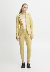 Žlutý přiléhavý blazer s odpovídajícími jogger kalhotami, doplněný bílým tričkem. Černé dřeváky dotvářejí celý outfit. Hladká látka, jednoduchý design.