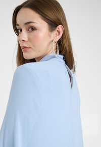 Světle modrá blůza s jemnou texturou, s vysokým límcem a výřezem vzadu. Velké, zdobené kruhové náušnice jsou viditelné.