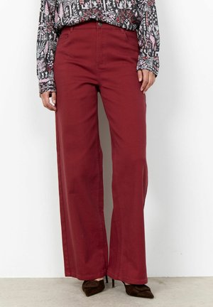 Persoon die bordeauxrode hoge taille wijde broek draagt, een blouse met lange mouwen en patroon, en donkerbruine puntige hakken, staand tegen een witte muur.