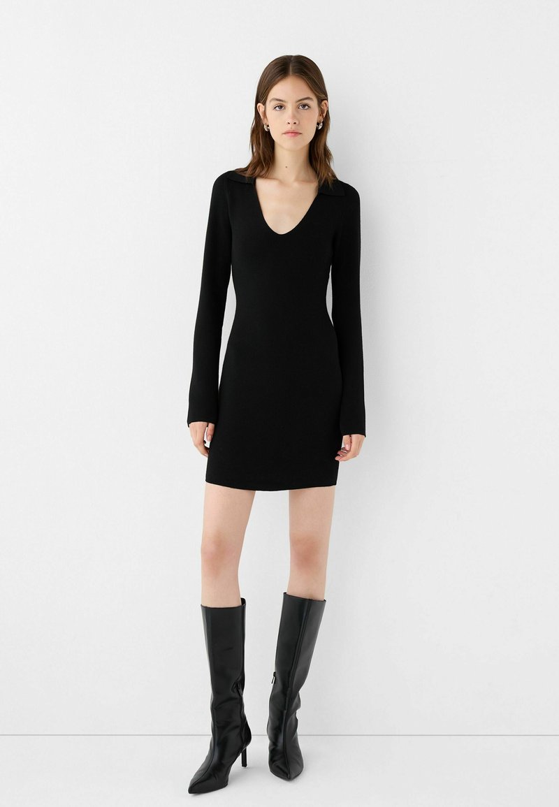 Abito mini nero a maniche lunghe con scollatura a V profonda, forma aderente e texture morbida, abbinato a stivali neri eleganti fino al ginocchio.