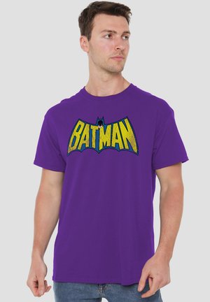 Mann trägt ein lila T-Shirt mit gelbem Batman-Logo und kleinem Batman-Kopf auf einfachem hellem Hintergrund.