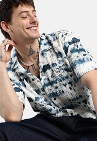 Camicia a maniche corte con bottoni, di colore bianco con motivi astratti blu e neri. Presenta una texture a coste e una vestibilità rilassata.