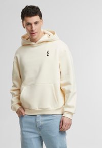 Kremfarget hettegenser med frontlomme, ribbestrikkede mansjetter og brodert "K" med et spadesymbol på brystet, sammen med lyseblå jeans.