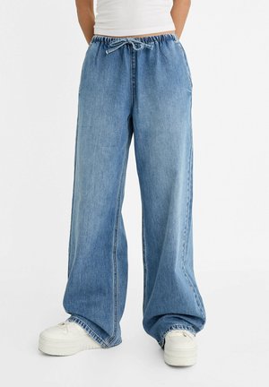 Person wearing loose blue denim pants with an elastic waistband and drawstring, paired with white platform sneakers.