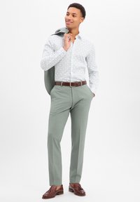 Camicia da uomo azzurra chiara con motivo, pantaloni verdi e mocassini marroni. L'outfit include una giacca grigia appoggiata sulla spalla.