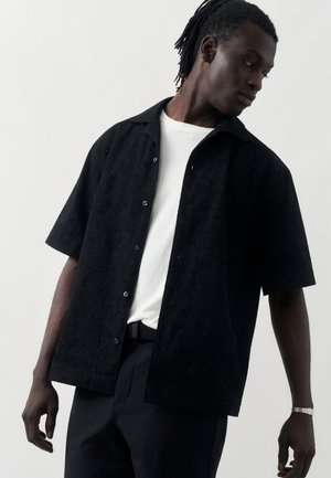 Černoch nosiaci textúrovanú čiernu krátku košeľu na gombíky cez biele tričko, čierne nohavice a náramkové hodinky, pozerá nabok.