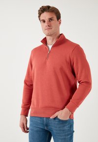 Sudadera en color coral, con un alto cuello con cremallera, puños de canalé y una textura suave. Combinada con vaqueros azules.