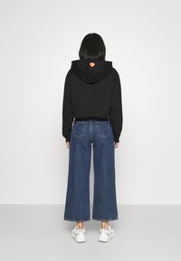 Hoodie noir court avec un détail en forme de cœur orange au dos, associé à un jean bleu à jambes larges et des baskets blanches. Texture de tissu lisse et décontractée.