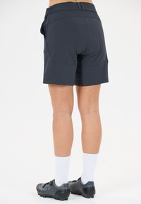 Person som bär svarta cykelshorts, vita strumpor och svarta cykelskor och står mot en enkel bakgrund.