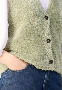 Fuzzy, lichtgroene vest met een V-hals ontwerp, voorzien van drie donkere knopen en een zak, gecombineerd met een donkere denim spijkerbroek.