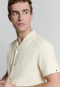 Korte mouwen, crèmekleurig shirt met een textuurstof, knoopsluiting aan de voorkant en een klein embleem op de mouw.