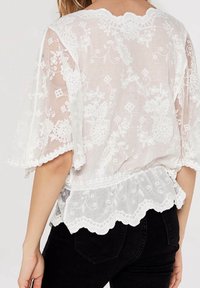Blusa blanca bordada con mangas cortas con borde en forma de ondas. Presenta patrones florales intrincados, un acabado texturizado y un dobladillo fruncido en la cintura.