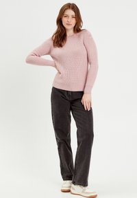 Geribbelde roze trui met lange mouwen, voorzien van knoopdetails op de schouder. Gecombineerd met donkergrijze jeans en crèmekleurige sneakers.