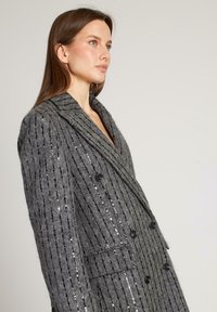 Abrigo gris de doble botonadura con rayas verticales, tejido texturizado y acentos de lentejuelas. Presenta un cuello de chal y dos bolsillos frontales.