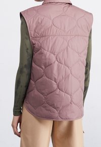 Pohodlná bezrukávová vesta v jemné fialkové barvě s prošívaným vlnitým vzorem, s límcem a měkkou, hladkou texturou, nošená přes tričko s dlouhým rukávem.