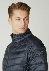 Jūrų laivyno pūkinė striukė su aukštu apykakle, su kvadratuotais raštais, užtrauktuku ir juodu logotipu kairiajame petyje. Minkšta, lengva medžiaga.