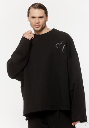 Mężczyzna o krótkich włosach, ubrany w oversize'ową czarną bluzę z czarną naszywką w kształcie nuty muzycznej na piersi, pozujący na białym tle.