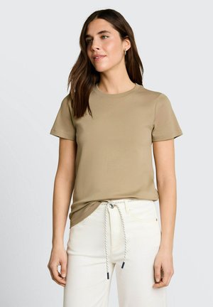 Femme aux longs cheveux bruns portant un haut beige à manches courtes et un pantalon blanc avec une ceinture à cordon nouée, debout devant un fond uni.